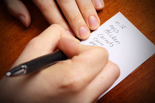 Close-up picture of person writing a grocery list that includes milk, eggs, chicken, carrots, corn, and peas