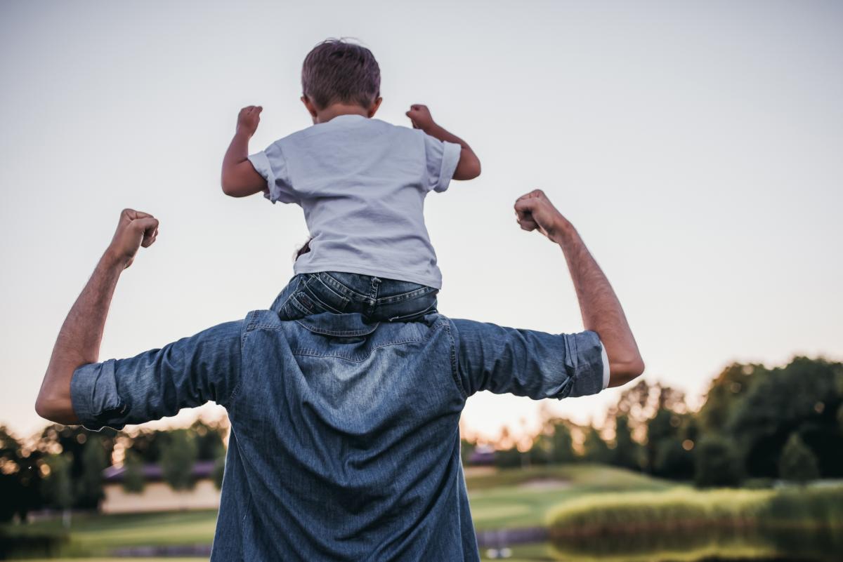 back view of a young boy on his dads shoulders playing outside. Both are flexing their arms.
