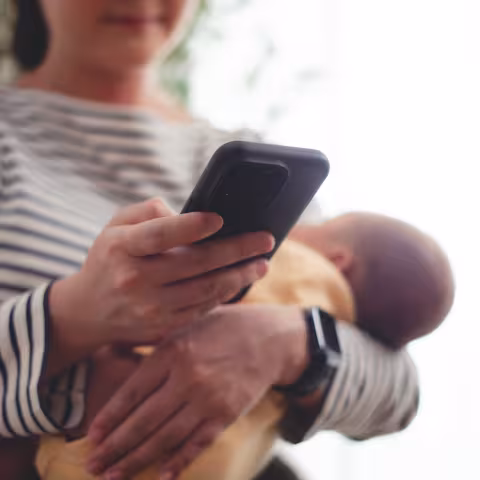 Close up shot of woman carrying her baby and taking a break using smartphone. 