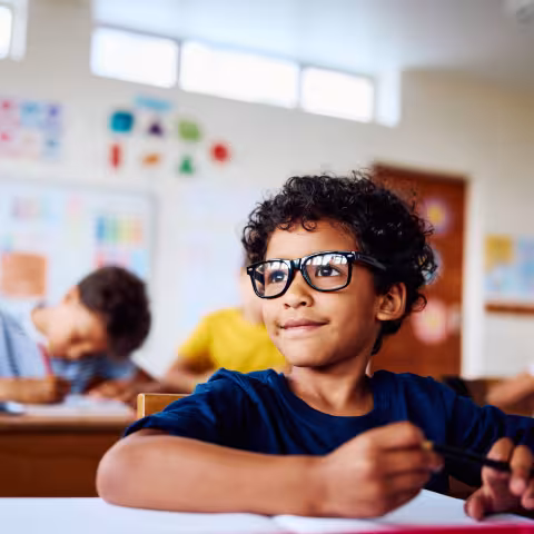 Schoolboy wearing eyeglasses sitting at desk, looking sideways, smiling in class