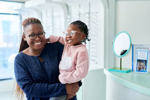Black woman holding her child, they are smiling wearing glasses in optometrist office 
