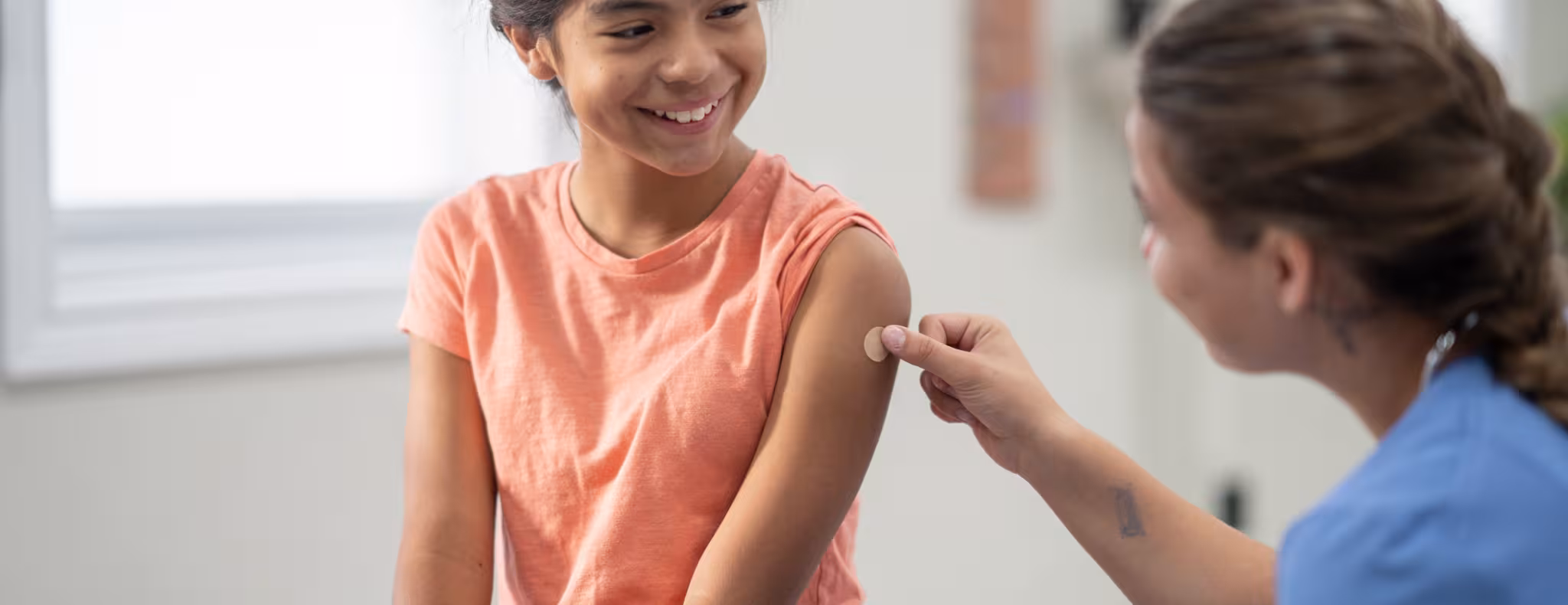 A school-aged girl sits up on an exam table as she holds still after a vaccination. She is dressed comfortably in a t-shirt and is smiling at her nurse as she places a bandage on the injection site.