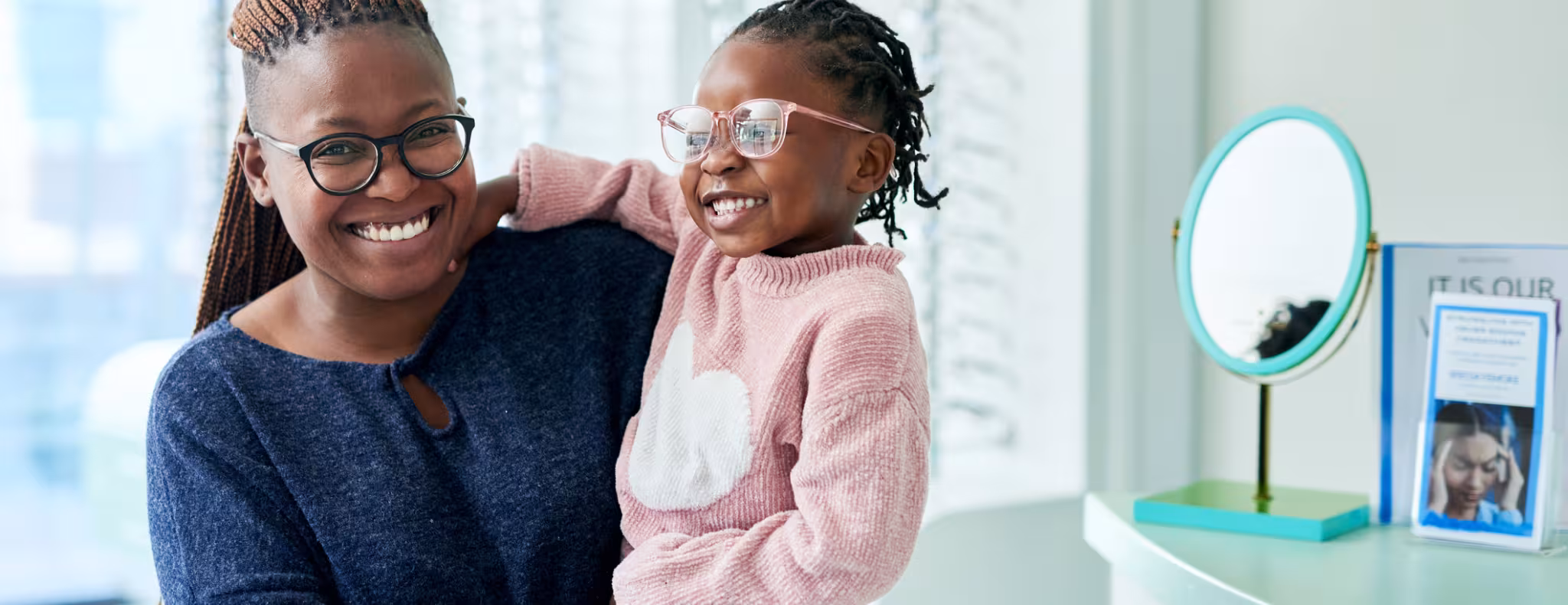 Black woman holding her child, they are smiling wearing glasses in optometrist office 